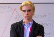 Young actor with blonde hair wearing a pinstriped suit and light blue shirt standing confidently with arms crossed in front of a whiteboard diagram