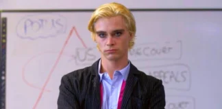 Young actor with blonde hair wearing a pinstriped suit and light blue shirt standing confidently with arms crossed in front of a whiteboard diagram