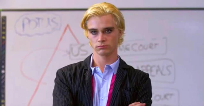 Young actor with blonde hair wearing a pinstriped suit and light blue shirt standing confidently with arms crossed in front of a whiteboard diagram