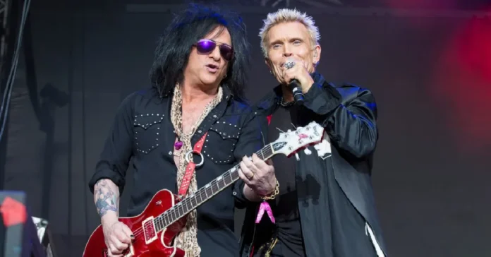 Steve Stevens performing live with Billy Idol holding red guitar on stage during concert showcasing his $45 million net worth career